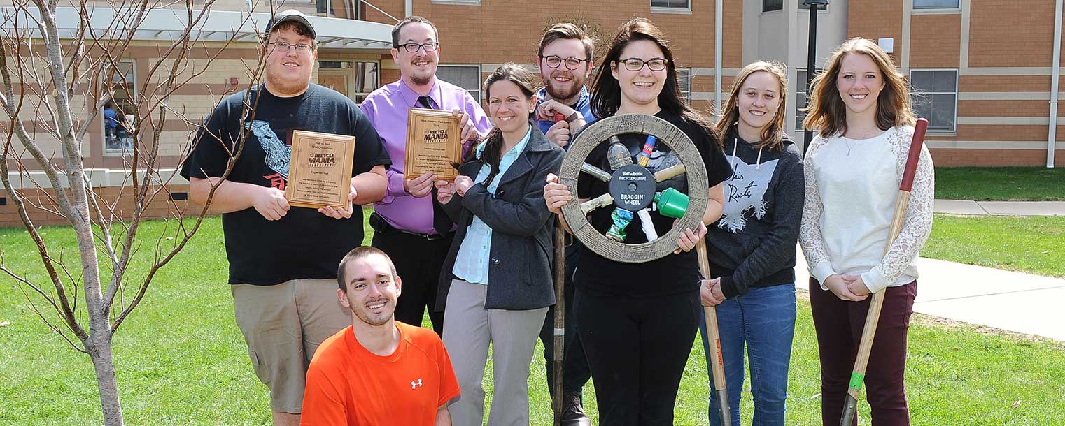 Kent State students show off their plaques from the residence hall challenge during this year’s RecycleMania recycling competition with Engleman Hall winning first place and Centennial A and B receiving “most improved.” The students also display the Braggin’ Wheel after defeating the University of Akron in the friendly environmental competition and stand next to the newly planted tree.